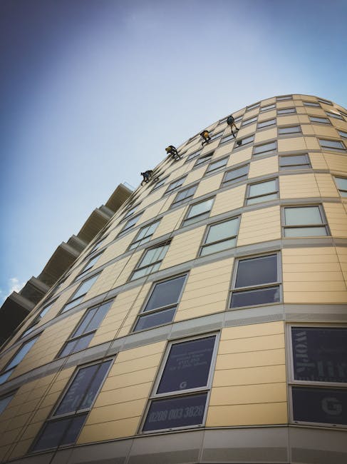 The image shows a modern, curved high-rise building with a facade composed of beige panels and numerous rectangular windows, some of which have reflective glass surfaces. The building appears well-mai