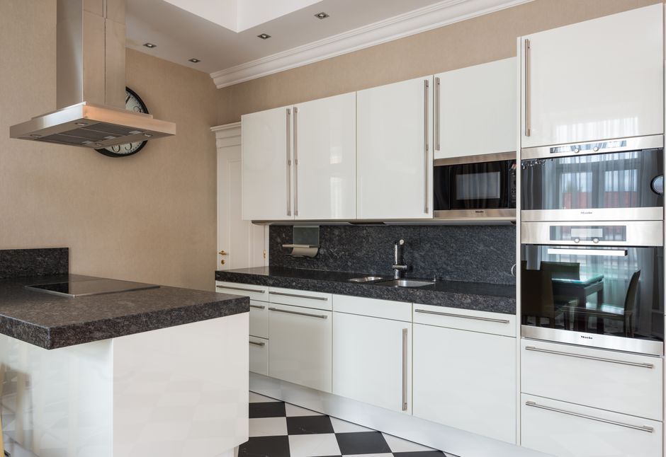 A person wearing yellow and blue rubber gloves is engaged in deep cleaning the interior of a white electric oven on a kitchen floor with black and white patterned tiles. The oven door is open, reveali